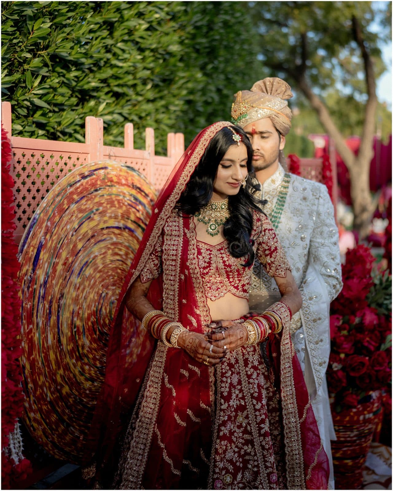 A bride and groom in traditional red and white attire beside a floral display
