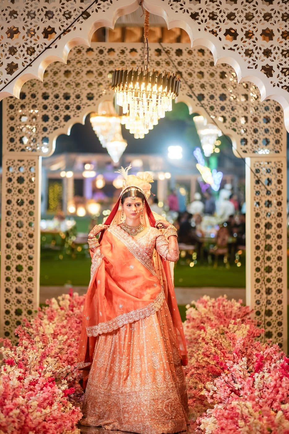 A bride in an orange lehenga under a carved archway